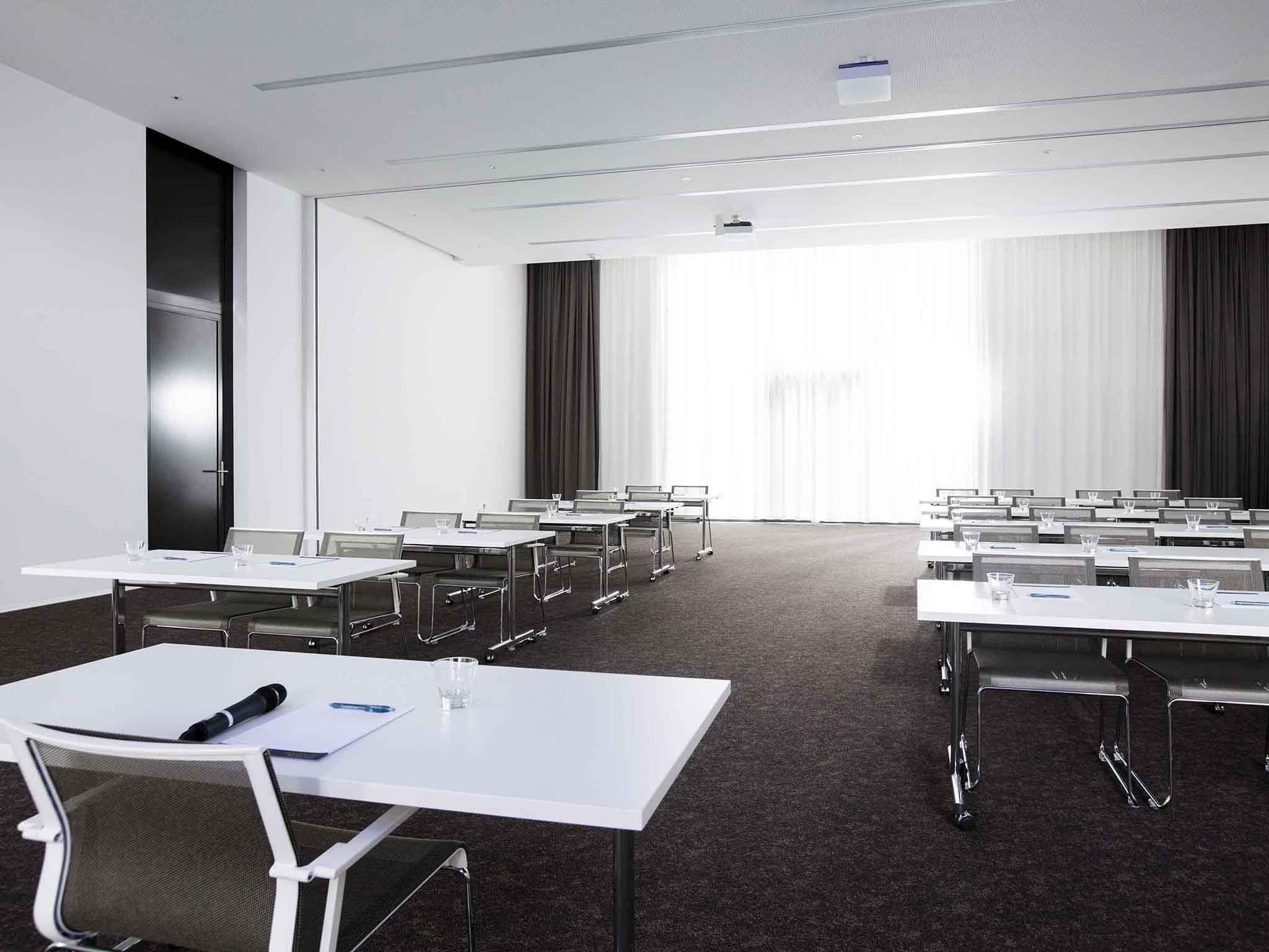 Une salle de conférence équipée de plusieurs tables, de chaises et d’une grande fenêtre avec des rideaux.