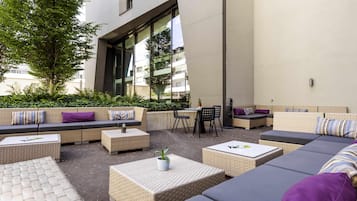 An outdoor seating area with sofas, chairs, and tables, surrounded by greenery and a building facade.