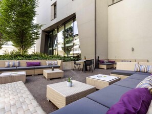 An outdoor seating area with sofas, chairs, and tables, surrounded by greenery and a building facade.