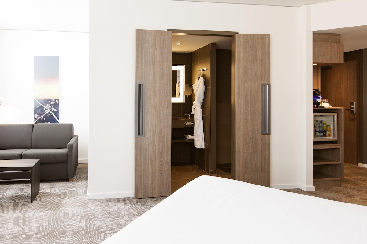 Habitación de hotel con cama, sofá gris, armario de madera y bata colgada en la puerta.