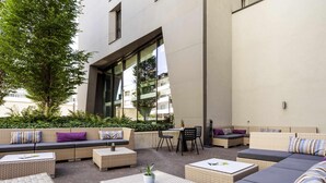 An outdoor seating area with sofas, chairs, and tables, surrounded by greenery and a building facade. - Novotel Basel City (Basel)