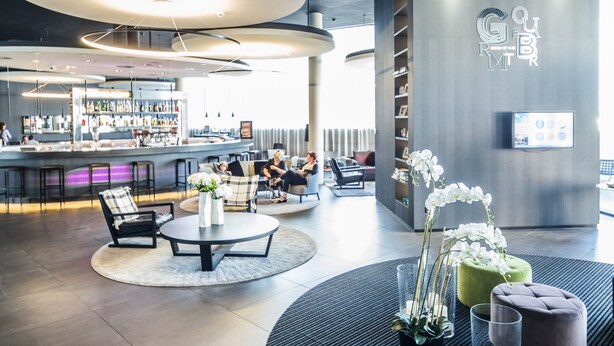 A modern lobby with circular ceiling lights, a bar area, seating arrangements, and a reception desk. - Novotel Basel City (Basel)