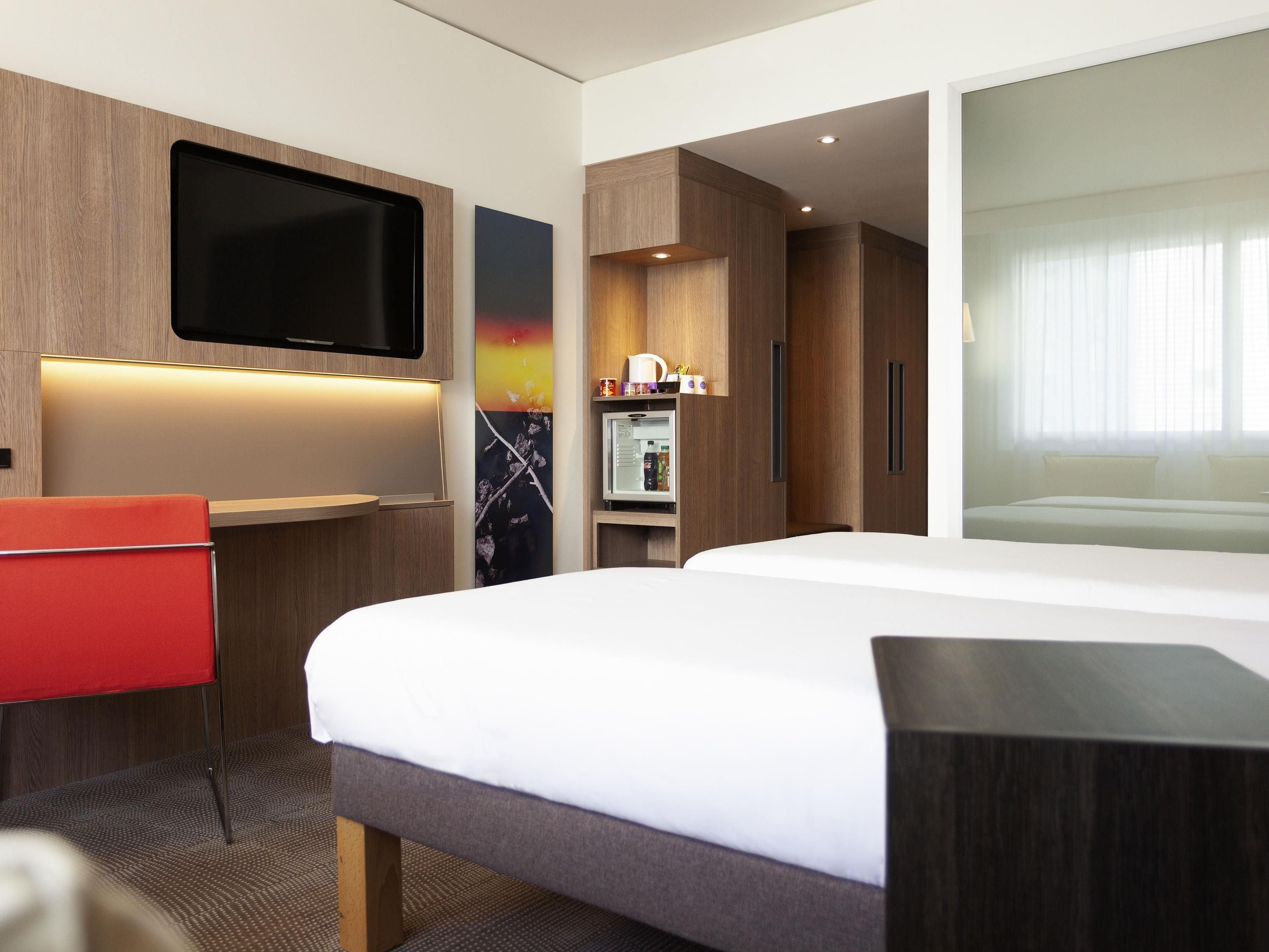 Quarto de hotel moderno com uma cama grande, televisão de tela plana na parede, uma pequena escrivaninha com cadeira e um refrigerador.