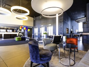 A modern hotel lobby with a reception desk, seating area, and a large circular light fixture. - Novotel Basel City (Basel)