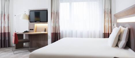 Habitación de hotel con cama, escritorio, silla, televisor y cortinas.