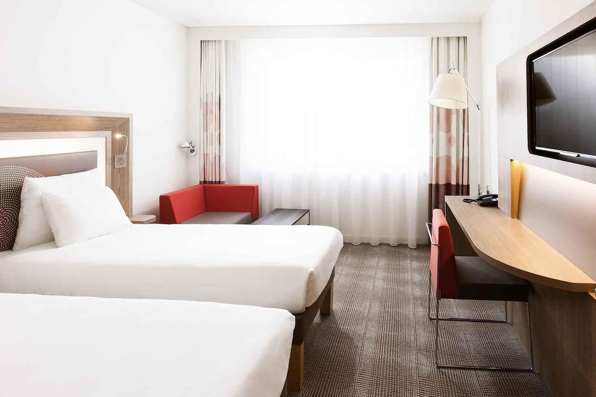 A hotel room with two beds, a desk, a TV, and a window with curtains.