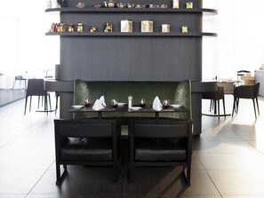 A dining area with a table set for two, a green upholstered bench, and a shelf above displaying various items. - Novotel Basel City (Basel)