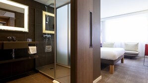 A modern hotel bathroom with a glass-enclosed shower, a vanity with a mirror, and a bed in the adjacent room. - Novotel Basel City (Basel)
