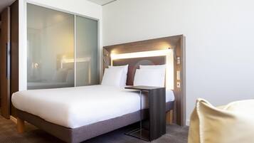 A modern hotel room with a large bed, a bedside table, a nightstand, and a glass partition wall.