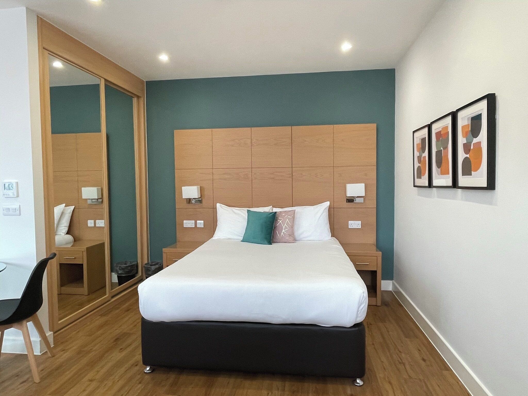 A modern hotel room with a large bed, wooden headboard, bedside tables, a desk, and a chair.