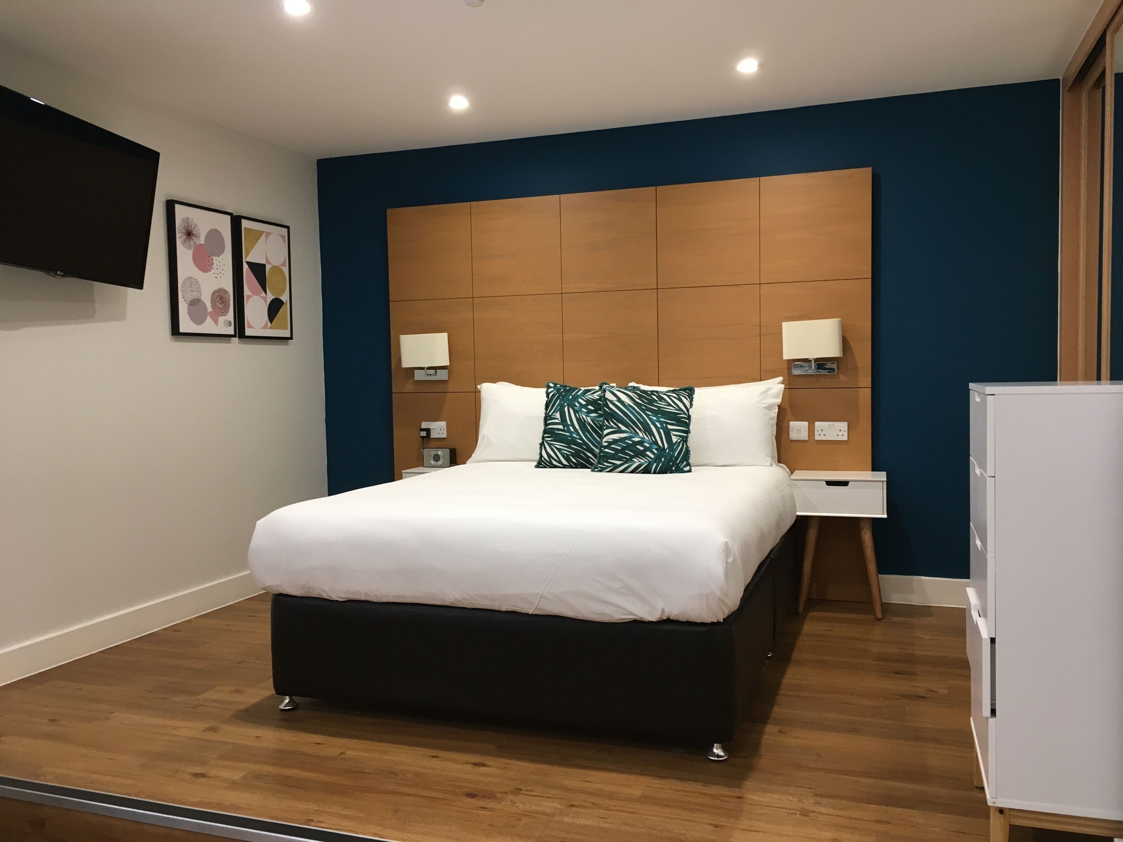 A modern hotel room with a large bed, wooden headboard, bedside lamps, and a flat-screen TV on the wall.