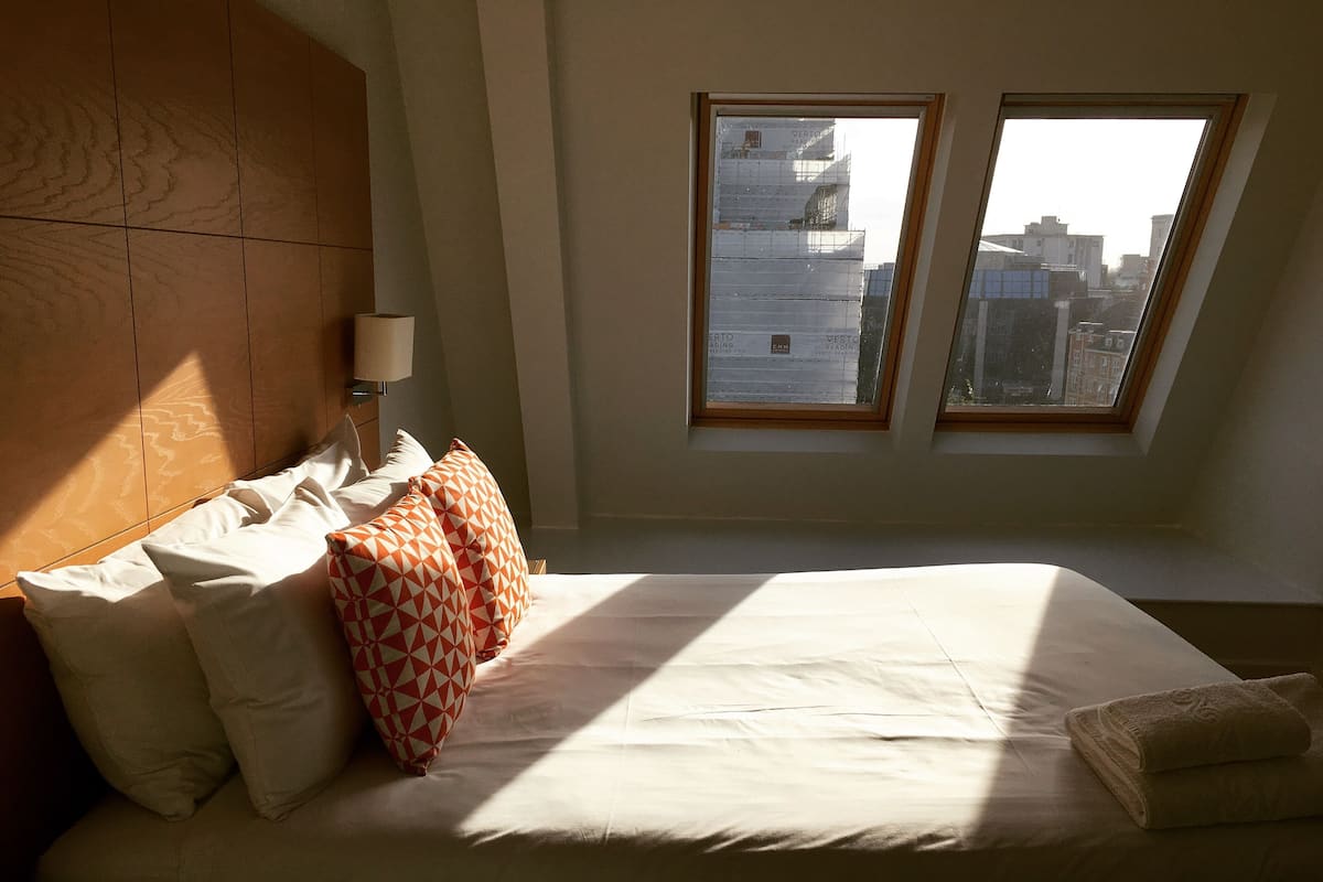 Une chambre d’hôtel avec un lit, des oreillers, une table de chevet et une fenêtre donnant sur des immeubles.