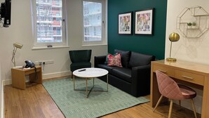 Een moderne woonkamer met een zwarte bank, een klein rond tafeltje en een tapijt met een groen patroon. Er zijn twee ramen met zonwering, een bureau met een stoel, en een wandplank met decoratieve objecten.