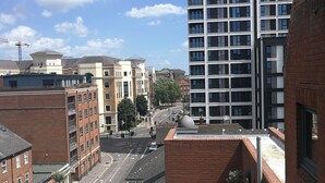 Een terras op het dak met buitenmeubilair, uitzicht op een stadsstraat en een hoog modern gebouw op de achtergrond.
