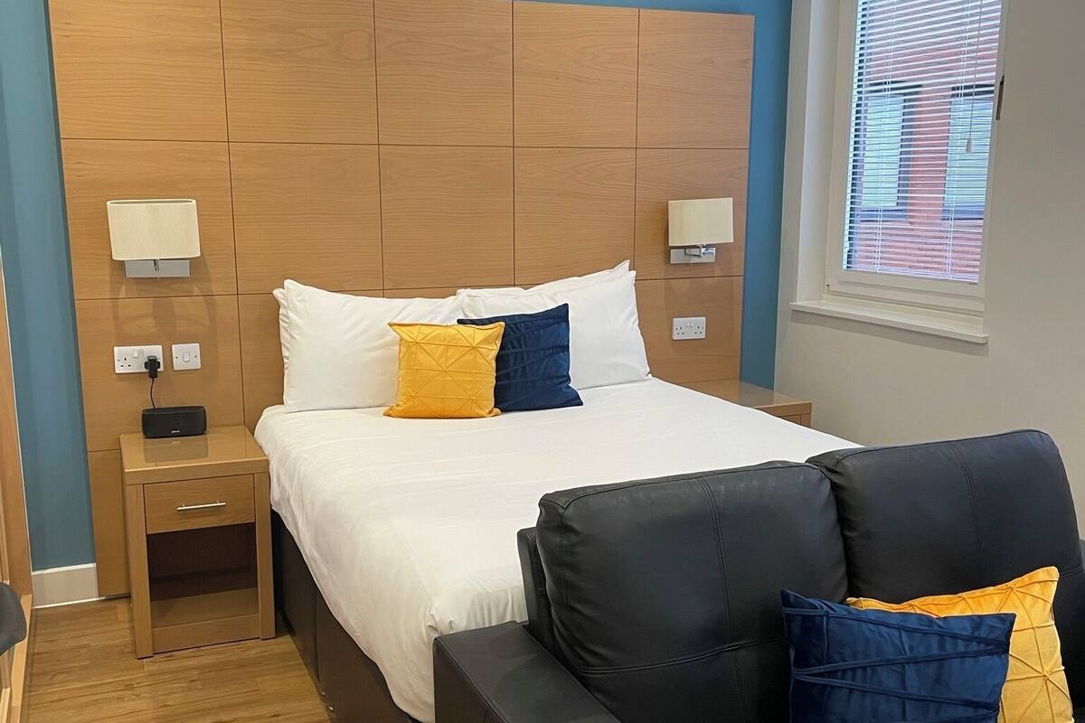 Une chambre d’hôtel avec une tête de lit en bois, un lit avec une literie blanche et bleue, une table de chevet avec une lampe, un canapé en cuir noir et une fenêtre avec des stores.