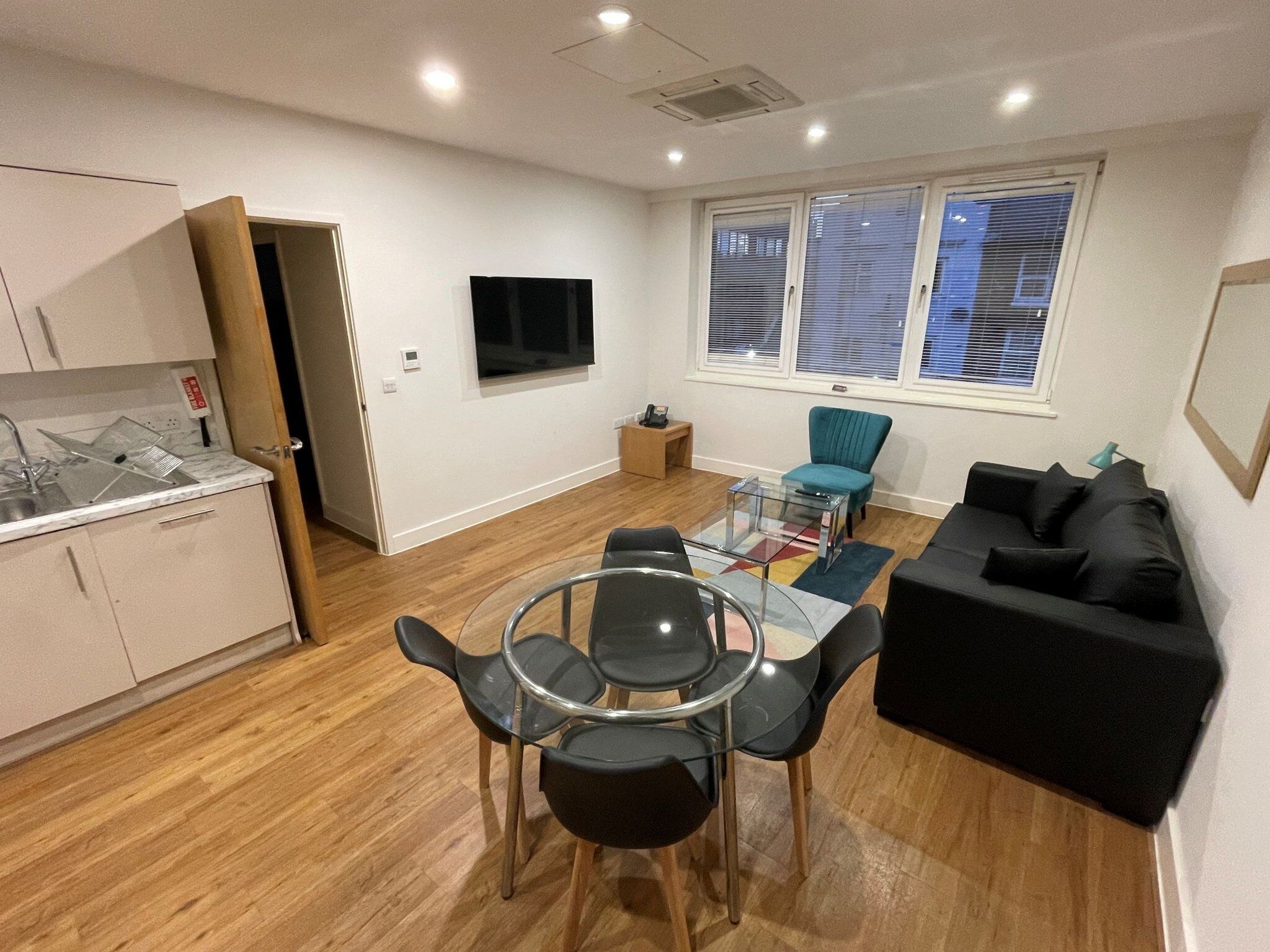 A compact living space with a kitchenette, a dining area with a glass table and chairs, a black sofa, and a TV mounted on the wall.