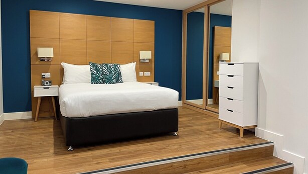 Een moderne hotelkamer met een groot bed, een houten hoofdbord, twee nachtkastjes met lampen, een garderobe en een spiegel.