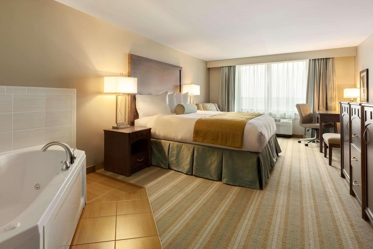 A hotel room with a large bed, a sitting area with a chair and table, a desk, a bathtub, and a large window with curtains.