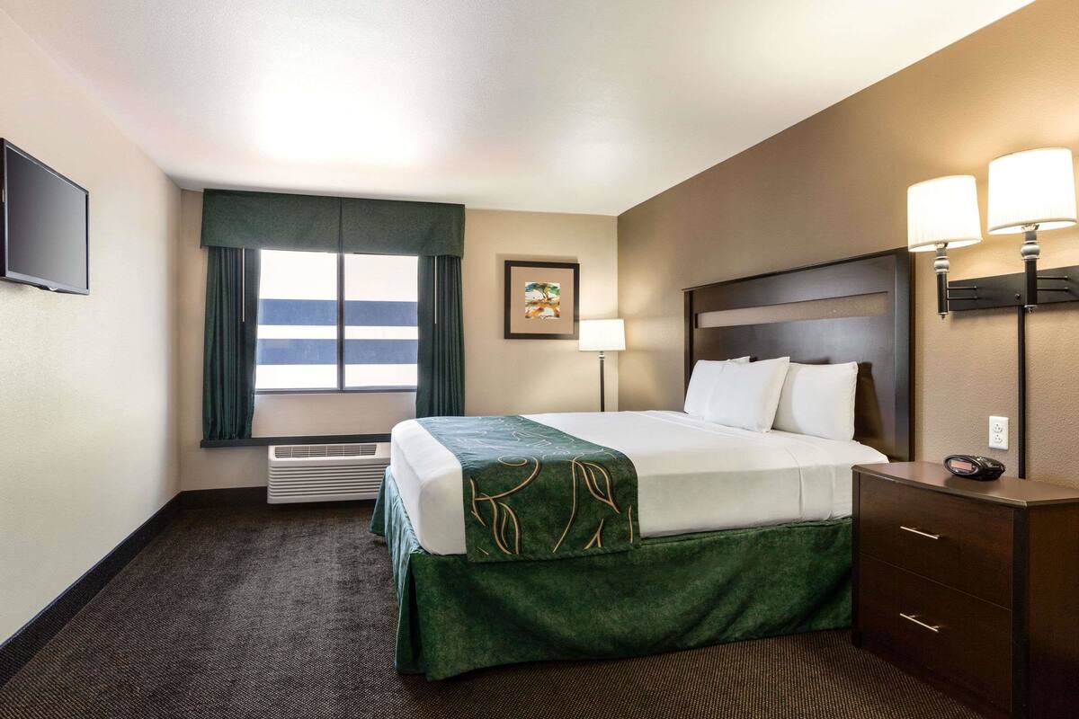 Ein Hotelzimmer mit Bett, Schreibtisch, Lampe, einem Bild an der Wand und einem Vorhangfenster.