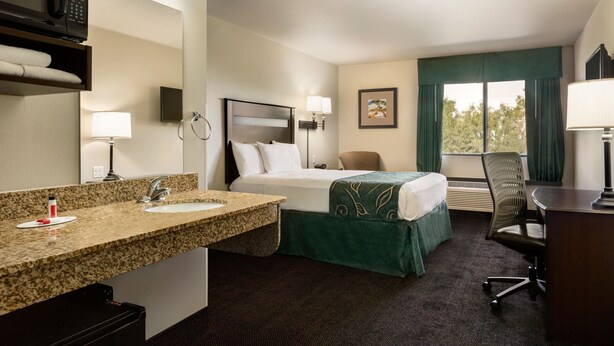 Ein Hotelzimmer mit Bett, Schreibtisch, Stuhl, Badezimmer mit Waschbecken und Spiegel sowie Fenster mit Vorhängen.