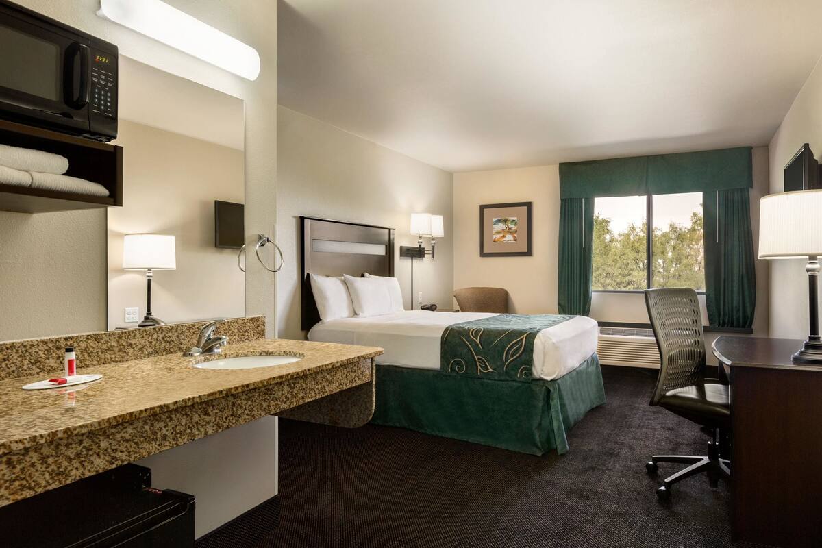 Ein Hotelzimmer mit Bett, Schreibtisch, Stuhl, Badezimmer mit Waschbecken und Spiegel sowie Fenster mit Vorhängen.