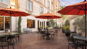 An outdoor patio area with tables and chairs, large potted plants, and red umbrellas. - Ayres Hotel Orange (Orange)