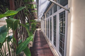 A narrow wooden pathway flanked by large green plants and a building with large windows. - The Sunrise Residence (Bangkok)