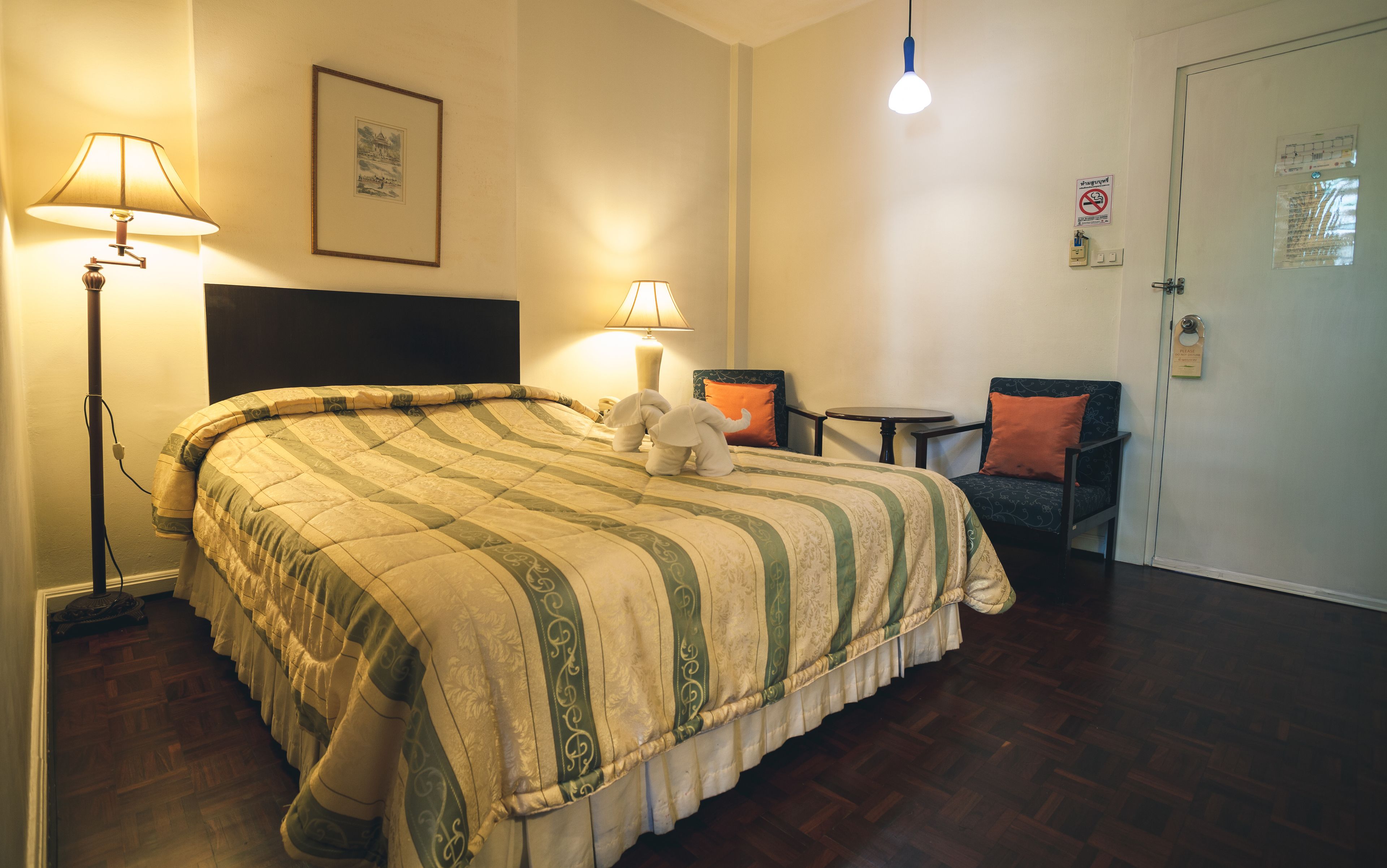A hotel room with a large bed, two bedside lamps, a small table, a chair, and a door with a sign.