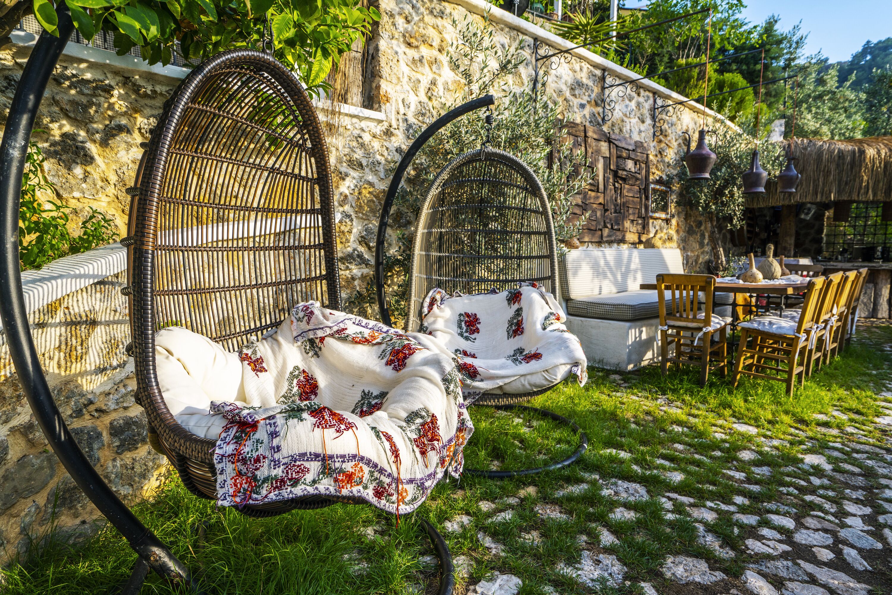 Una silla de mimbre con cojín floral, un sofá de mimbre y una mesa con sillas en un jardín con muros de piedra y follaje.