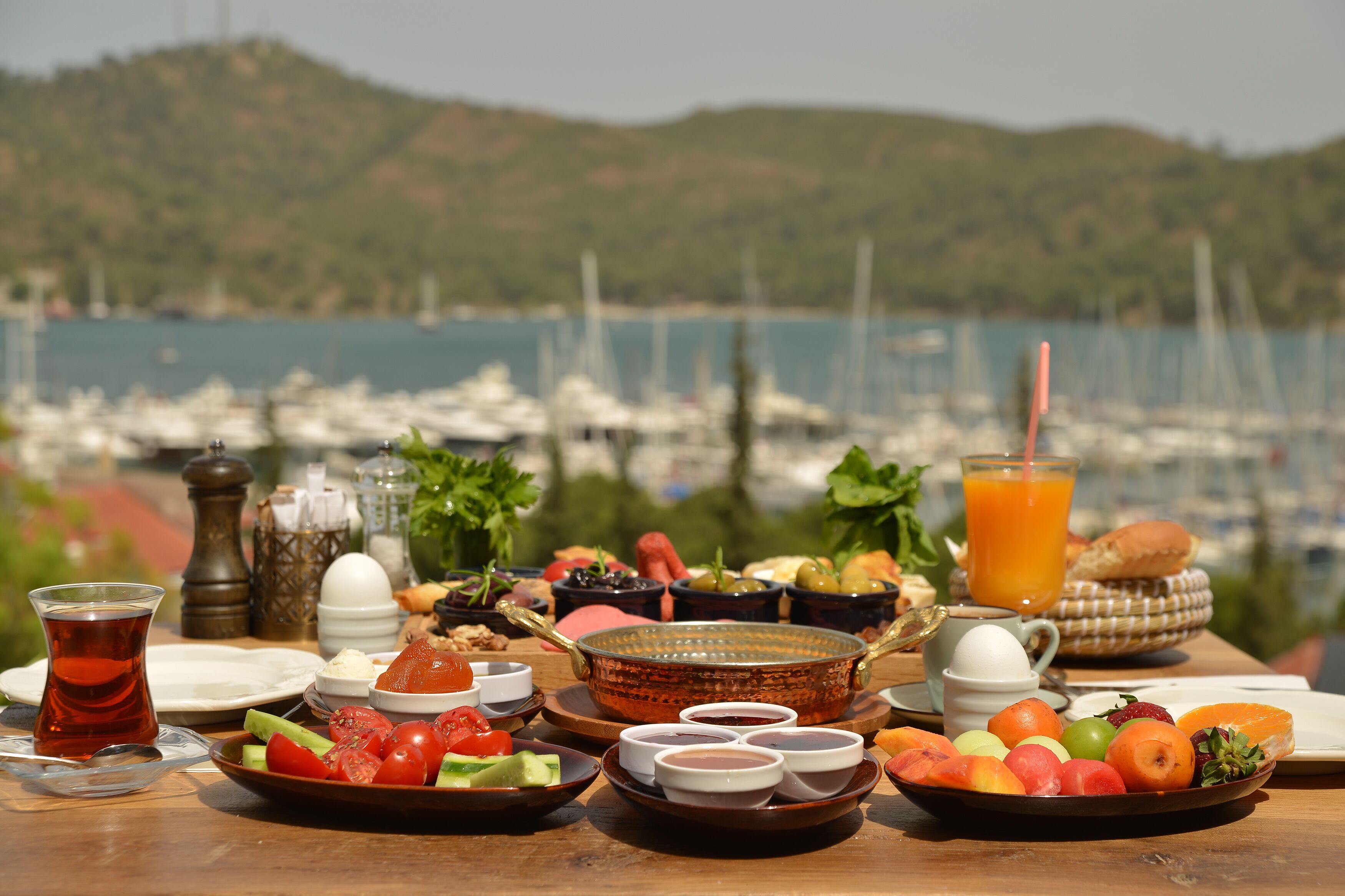 A table set with a variety of foods and drinks, including fruits, vegetables, and a glass of orange juice, with a scenic view of boats and hills in the background.