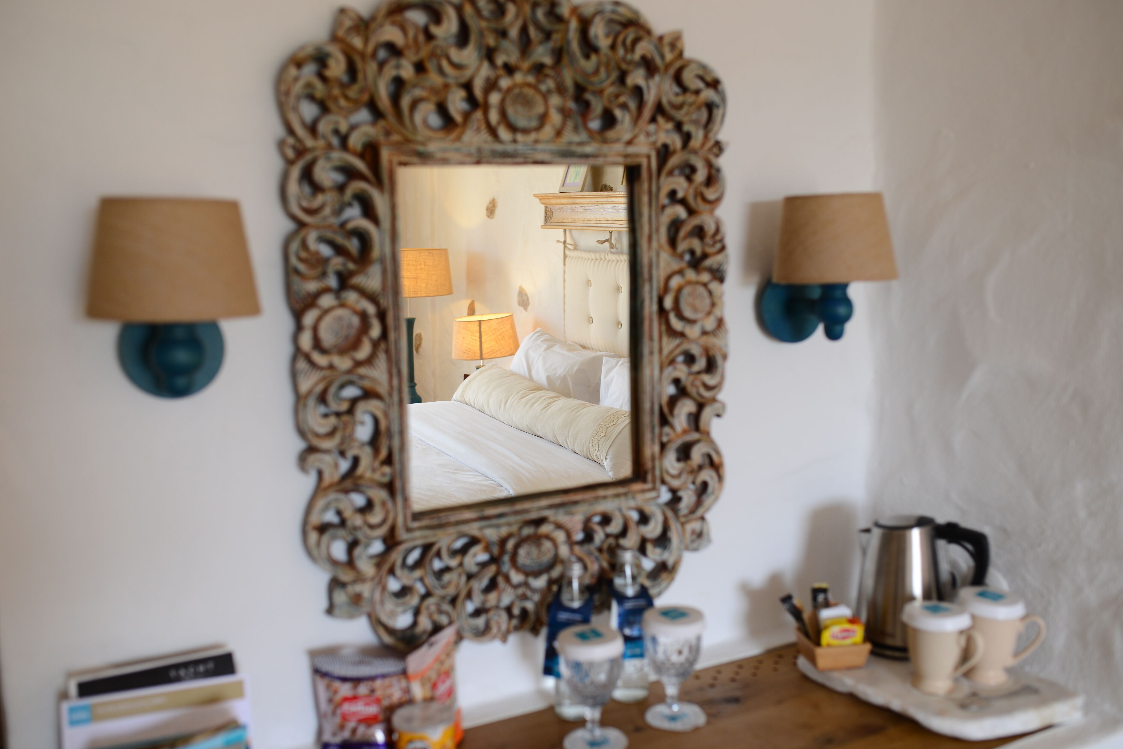 Een slaapkamer met een grote, versierde spiegel, twee wandlampen, een nachtkastje met waterkoker, kopjes en een klein kastje.