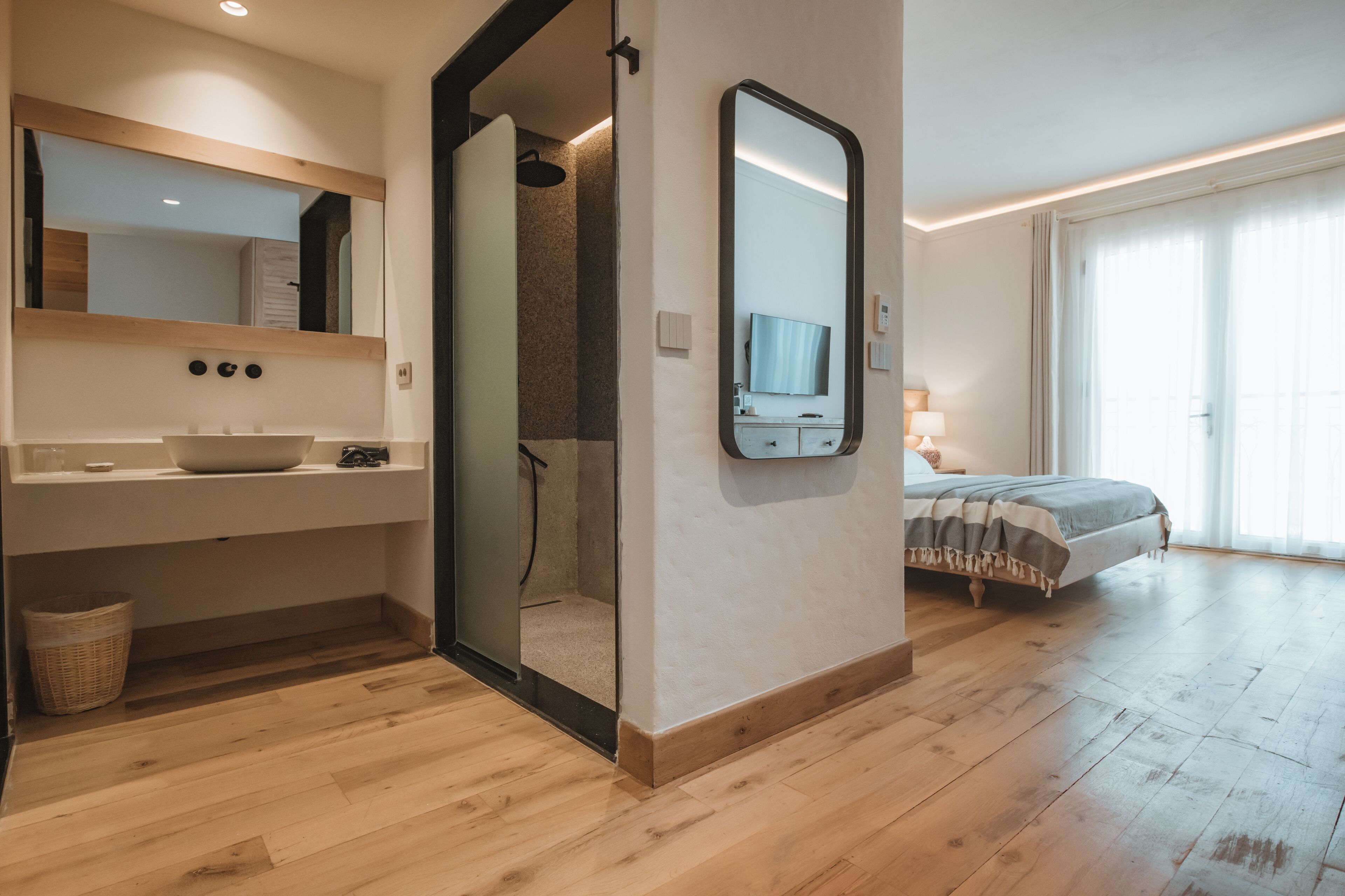 Una habitación de hotel moderna con piso de madera, un espejo grande, un lavamanos, ducha, cama y televisor.