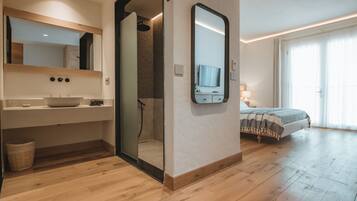 Una habitación de hotel moderna con piso de madera, un espejo grande, un lavamanos, ducha, cama y televisor.