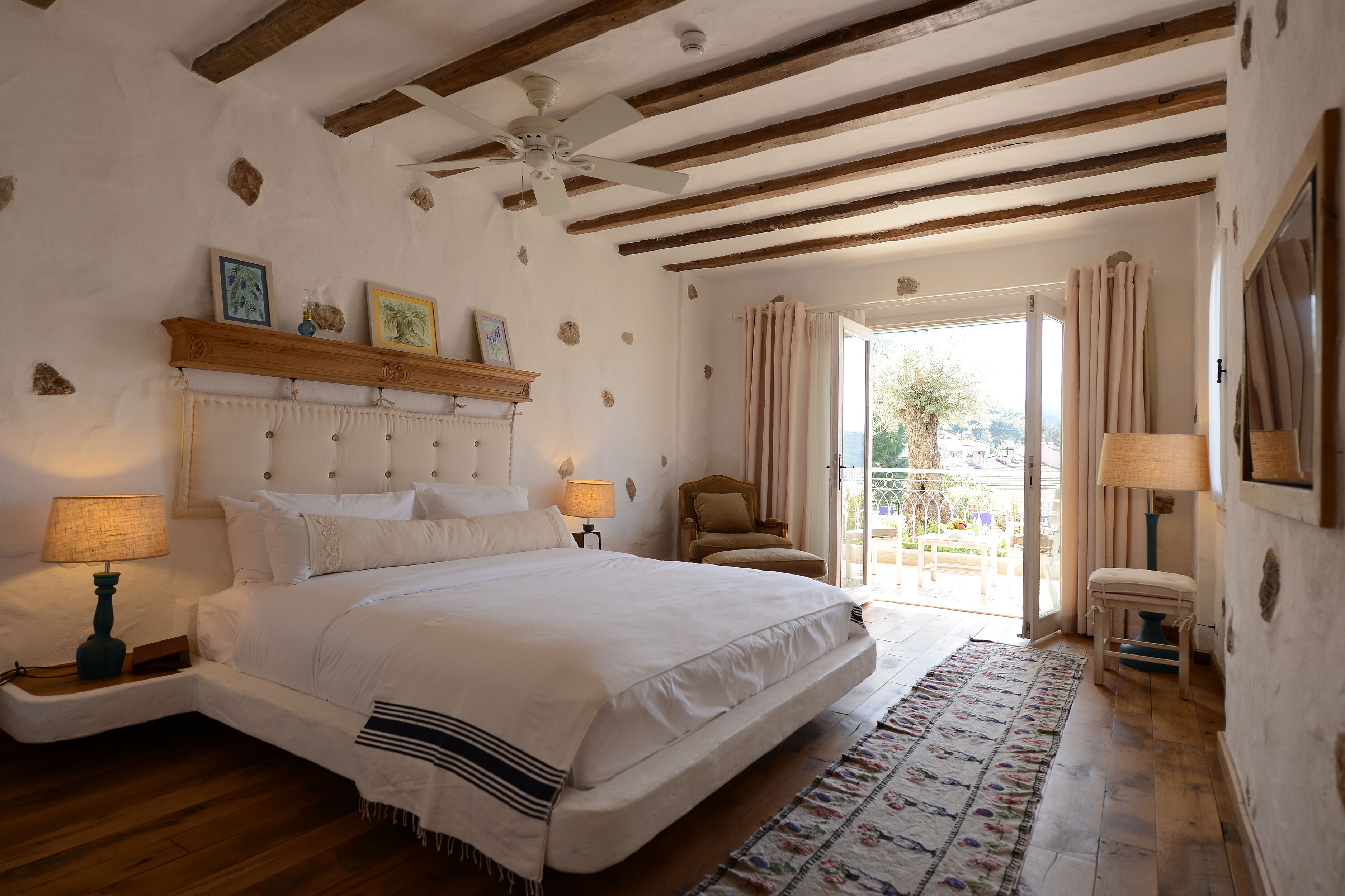 A bedroom with a large bed, wooden beams, a ceiling fan, a nightstand with a lamp, a chair, and a view through a glass door.