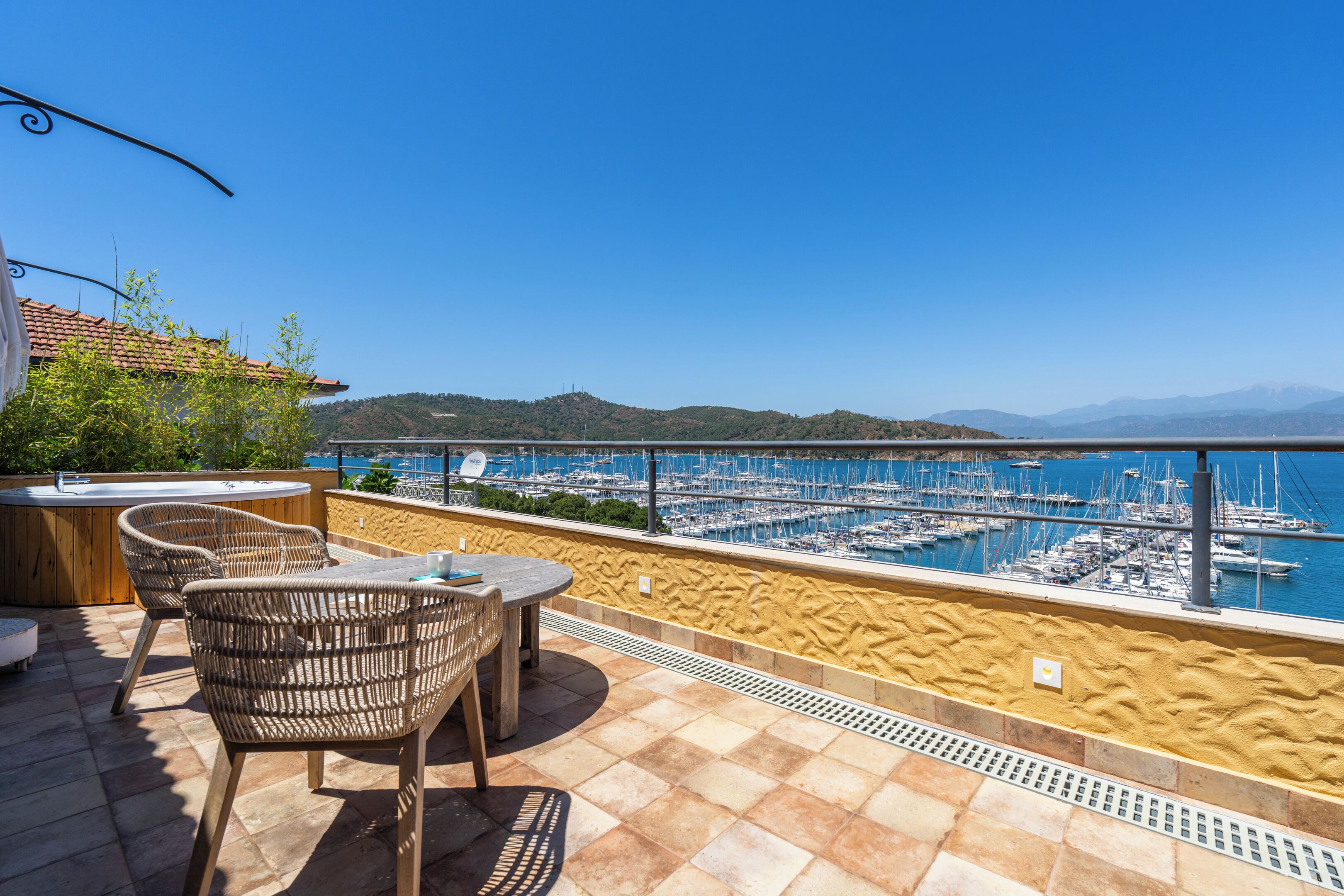 A terrace with wicker chairs and a table, overlooking a marina with boats and a hilly landscape.