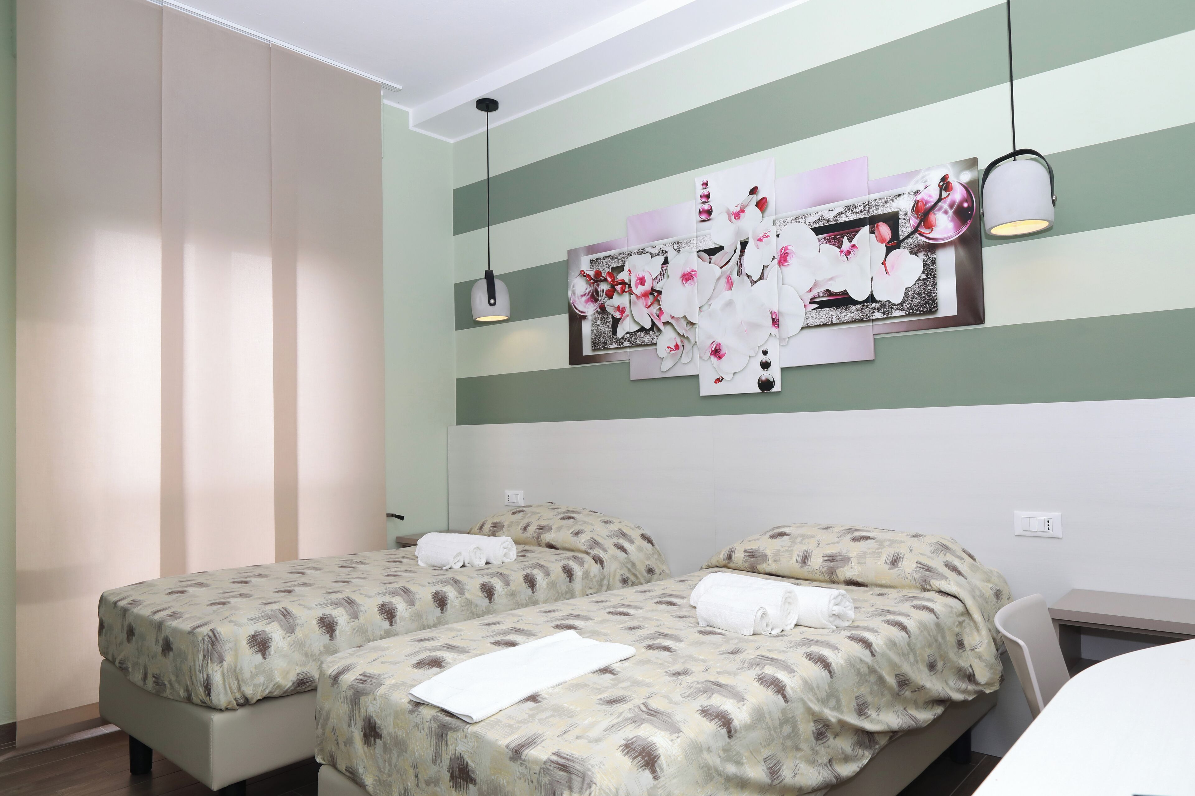 A hotel room with two beds, each with a patterned bedspread and a rolled towel. There is a wall-mounted artwork above the beds and a hanging pendant light.