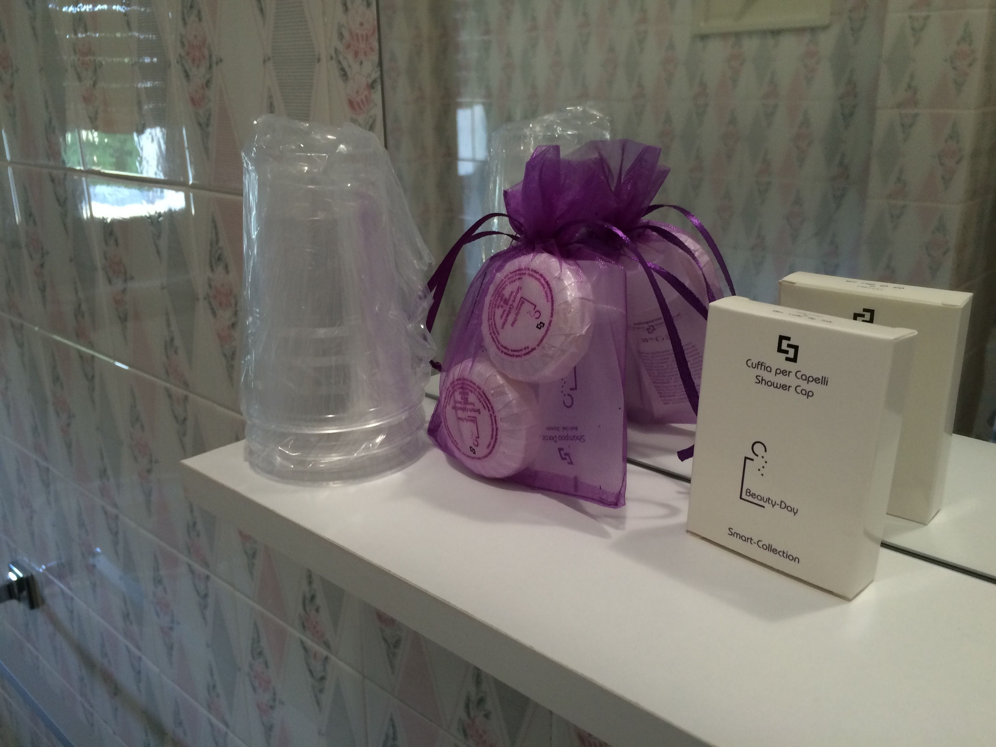 Un estante de baño con un gorro de ducha, una bolsa de regalo morada y una caja etiquetada "Cuffia per Capelli Shower Cap Beauty-Day Smart-Collection".