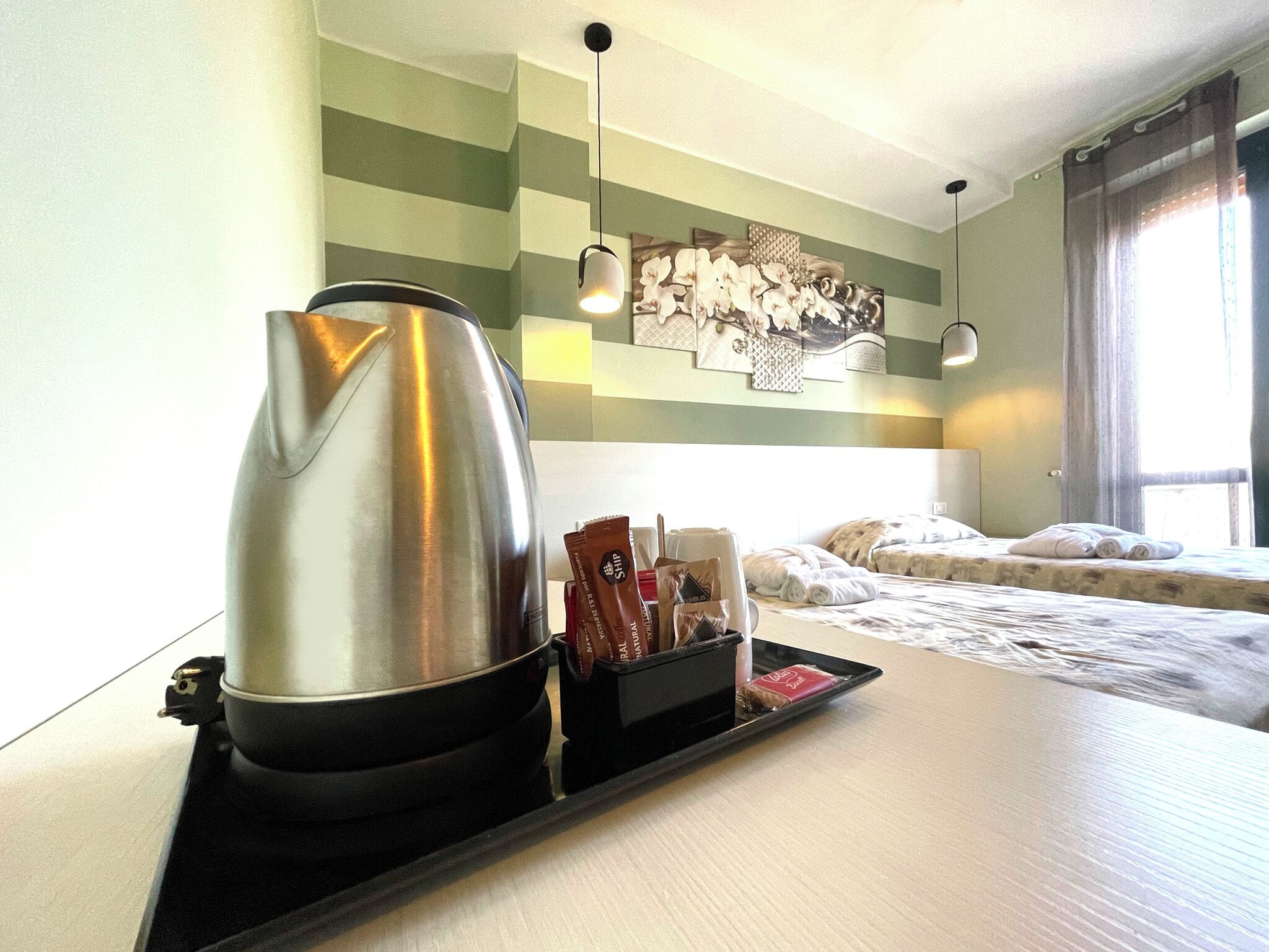 A hotel room with a bed, a tray with a kettle and toiletries, a wall with a patterned artwork, and a window with curtains.