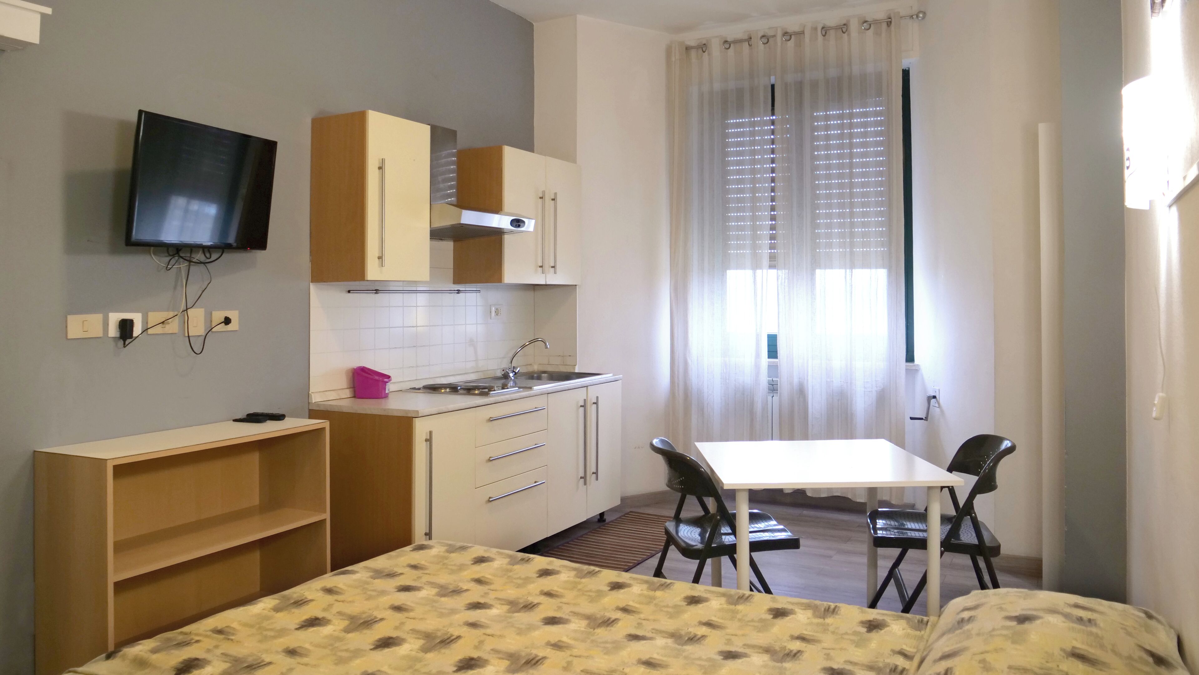 Quarto compacto com cama, pequeno espaço de cozinha, mesa de jantar com cadeiras e televisão na parede.