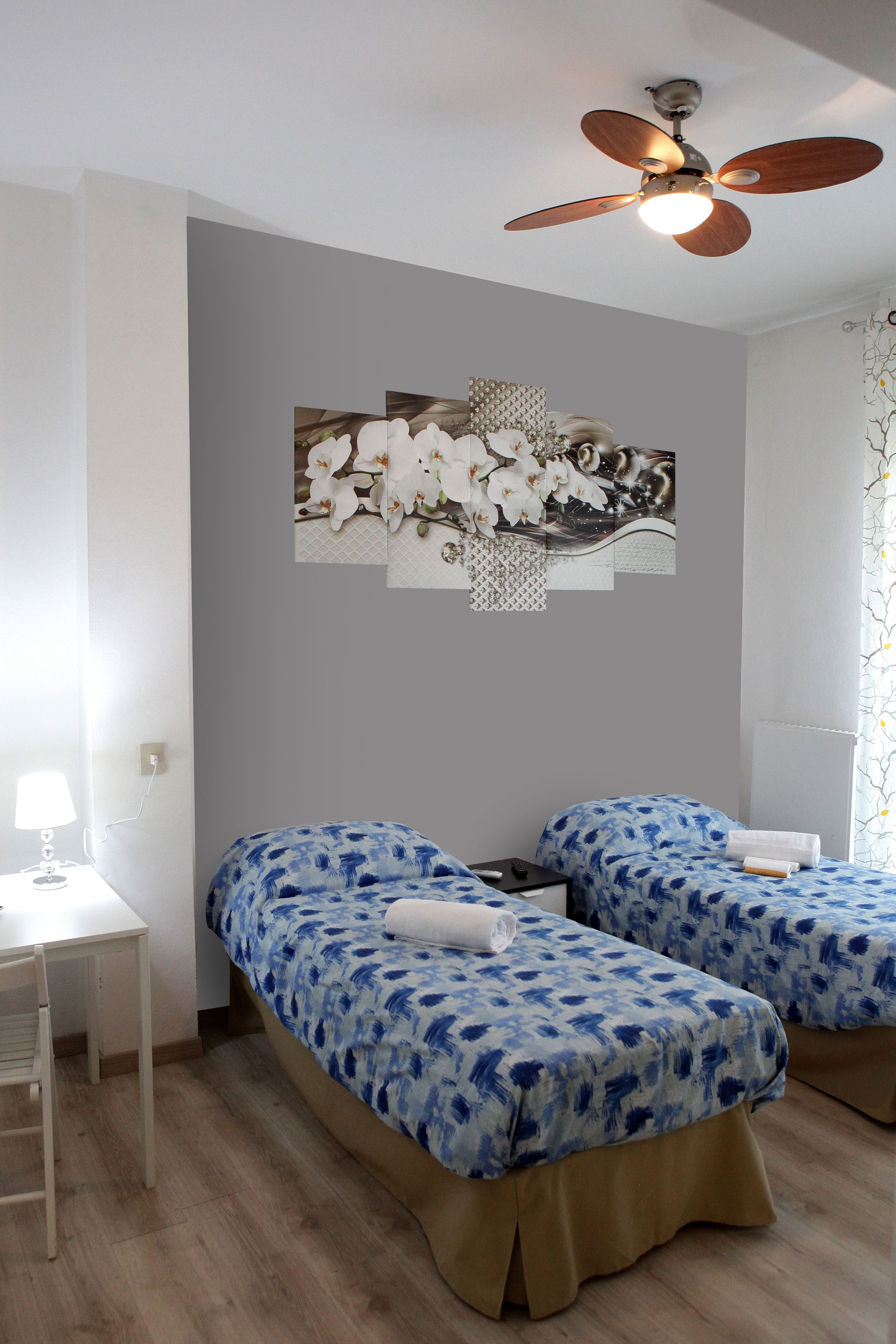 A room with two beds, a ceiling fan, a desk with a lamp, and a wall with a floral artwork.