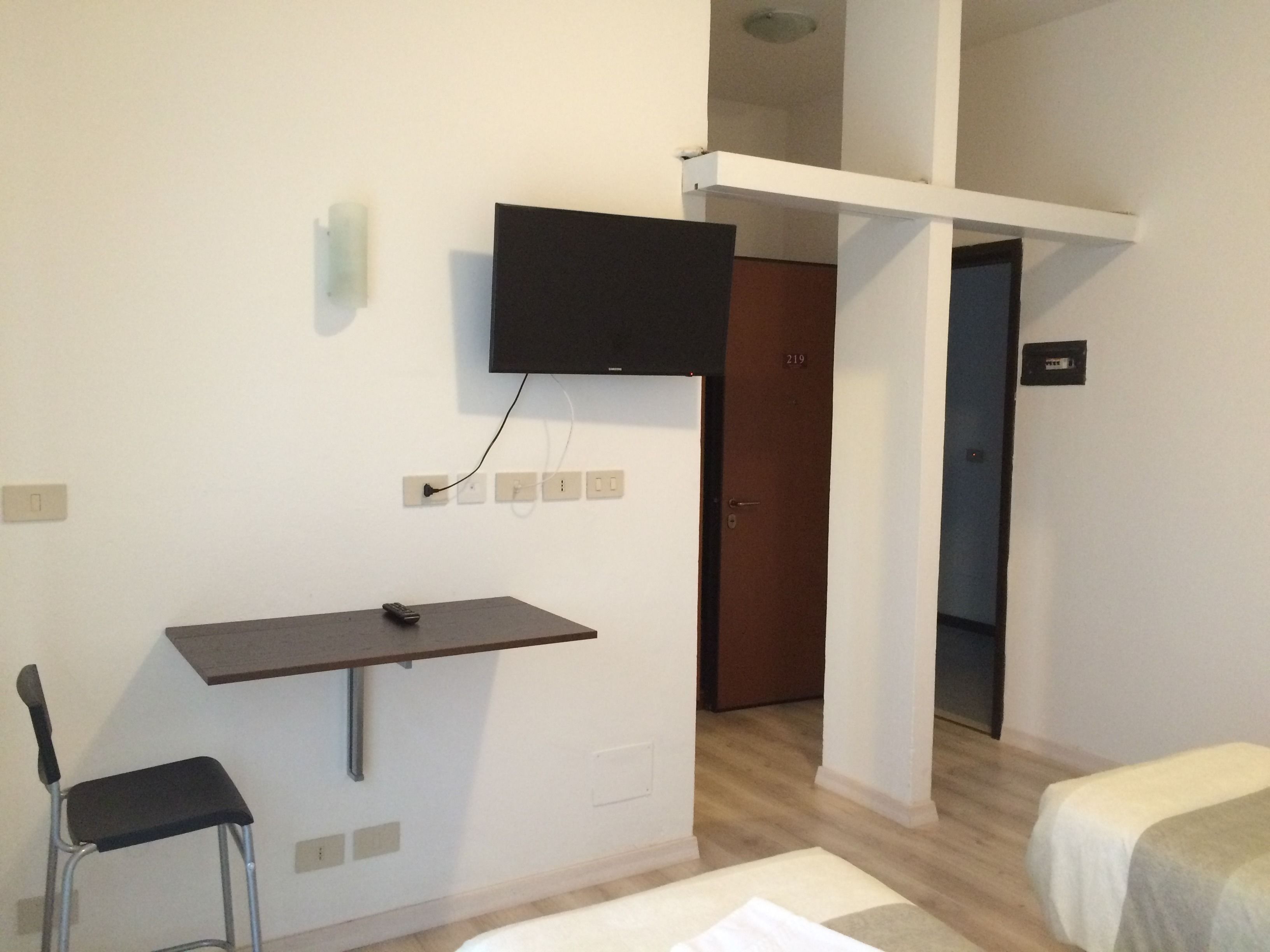 Een kleine, eenvoudig ingerichte kamer met een bureau, een stoel, een bed en een aan de muur bevestigde televisie.