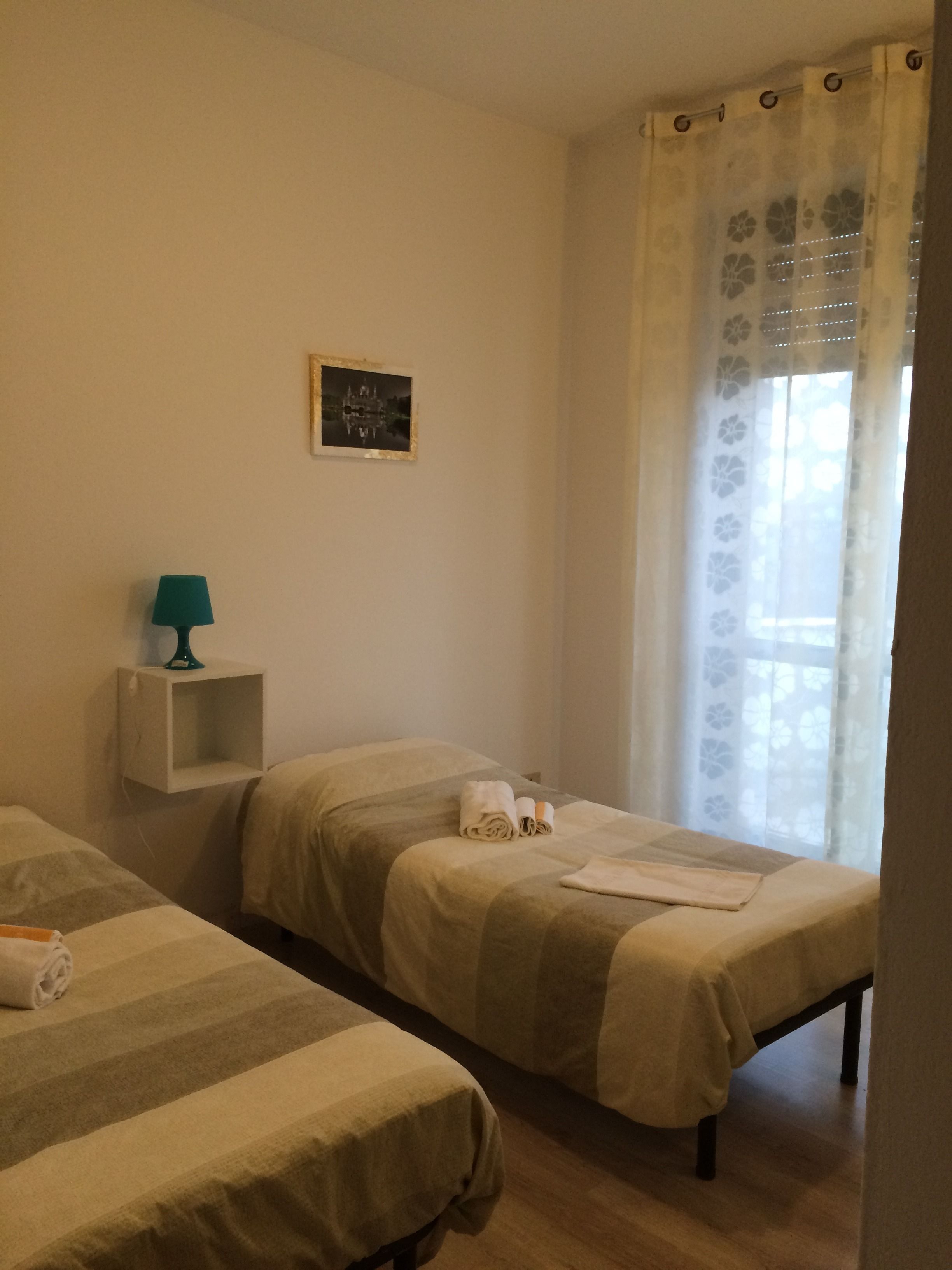 A room with two single beds, a small shelf with a lamp, a framed picture on the wall, and a window with a floral curtain.