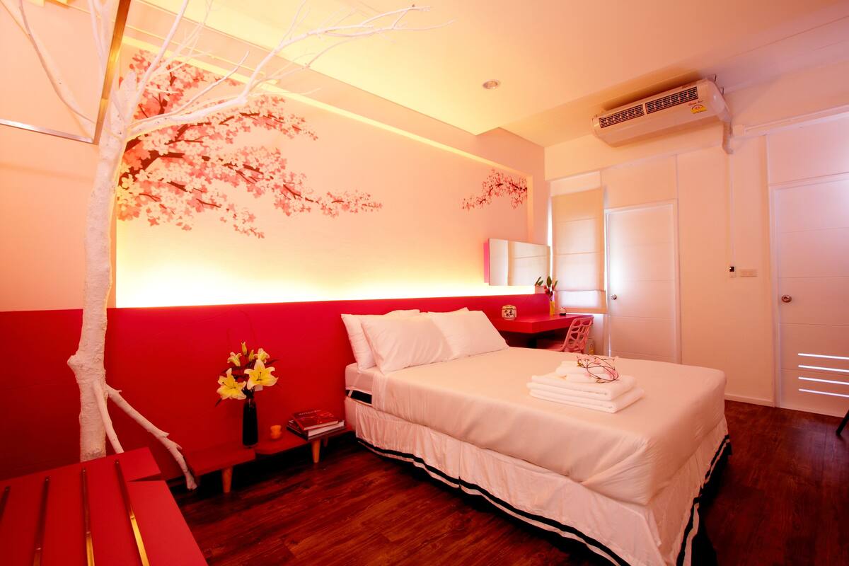 A hotel room with a bed, a nightstand, a vase with flowers, a red bench, and a wall with cherry blossom decorations.