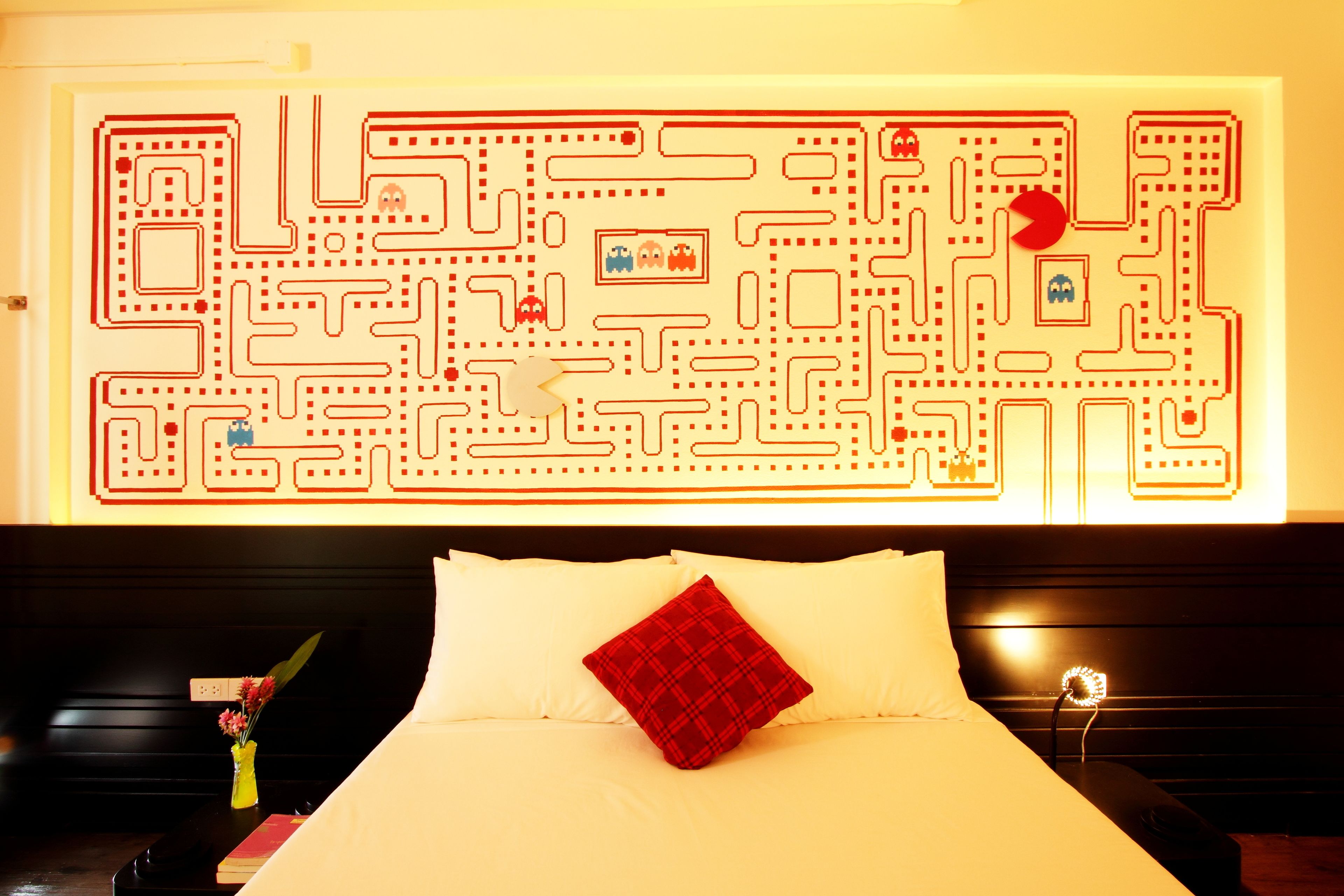 A hotel room with a maze-like wall art, a bed with white and red pillows, and a bedside table with a vase of flowers.