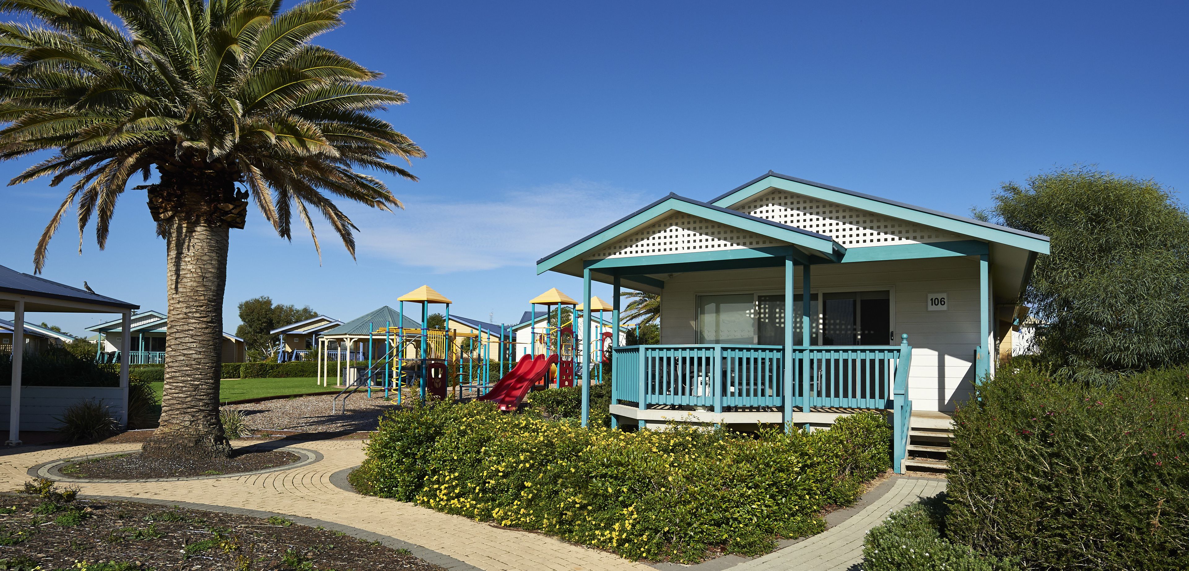A playground with slides and climbing structures, a palm tree, and a building with a porch.