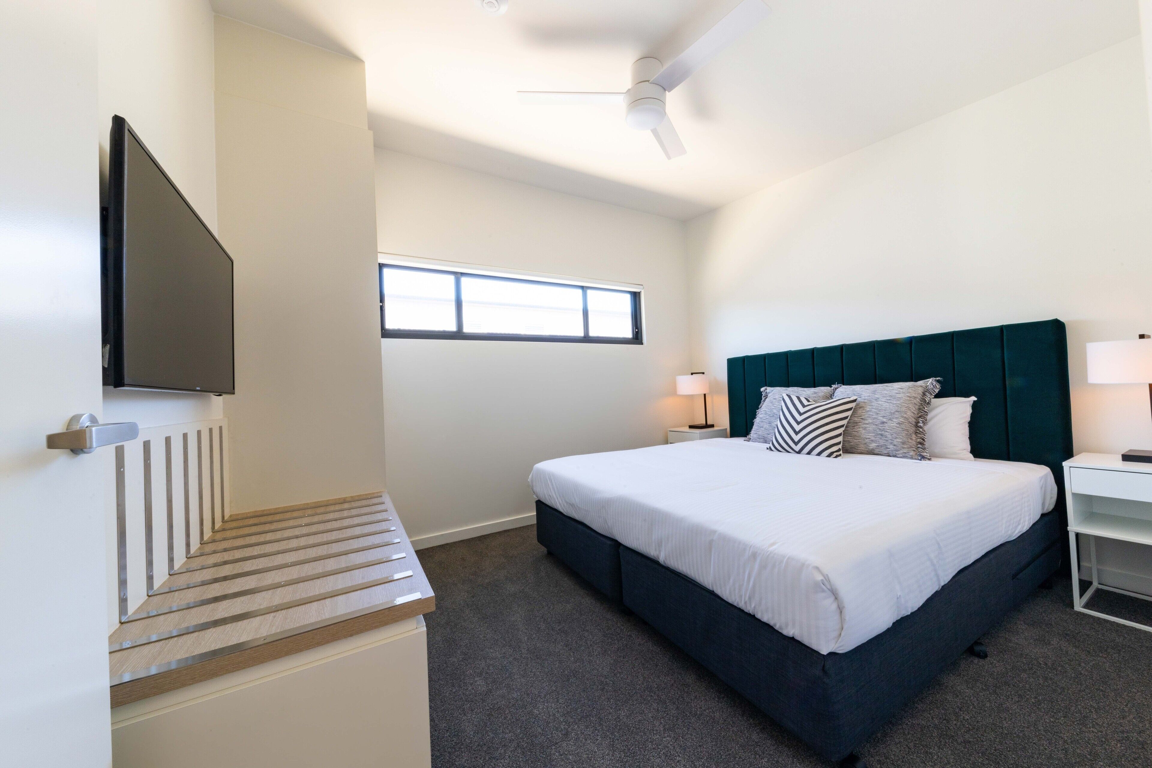A hotel room with a bed, a TV mounted on the wall, a window, and a ceiling fan.