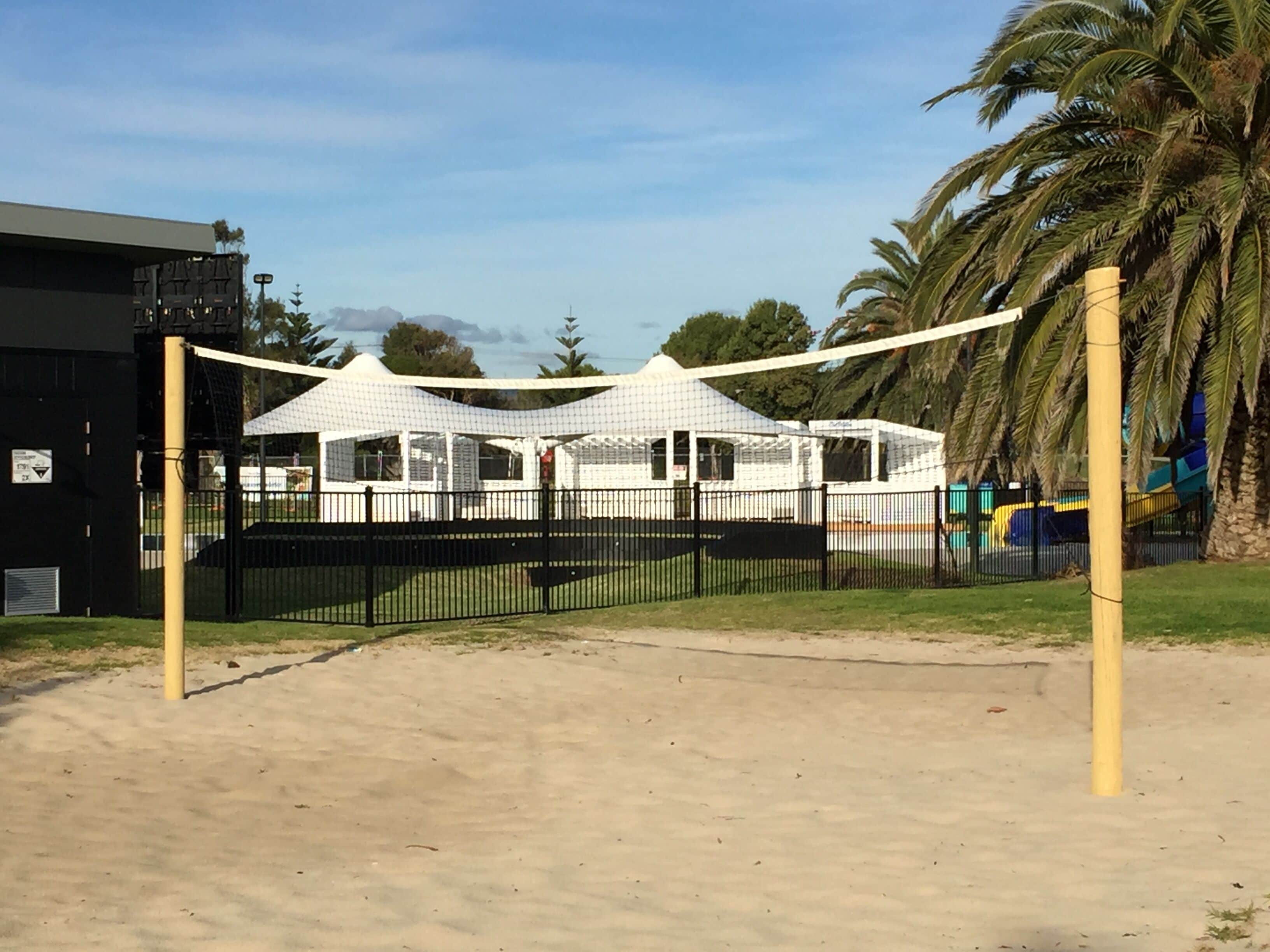 Un terrain de volley-ball de plage avec un filet, entouré d’une clôture noire et de palmiers.