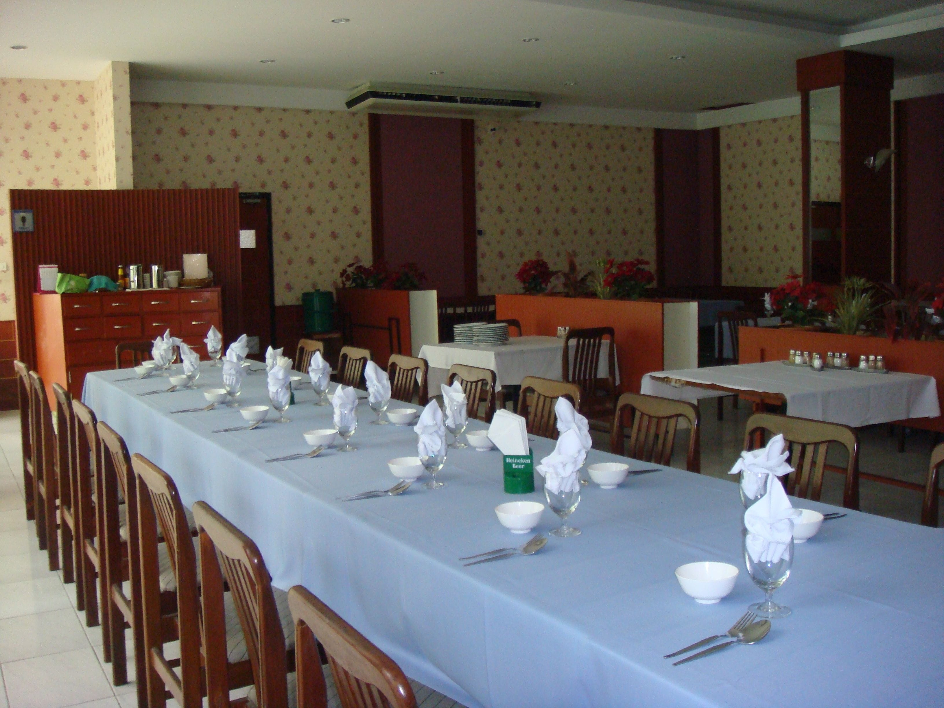 Un espace repas avec de longues tables dressées pour accueillir plusieurs convives, agrémenté de nappes blanches, de couverts en argent et de verres.