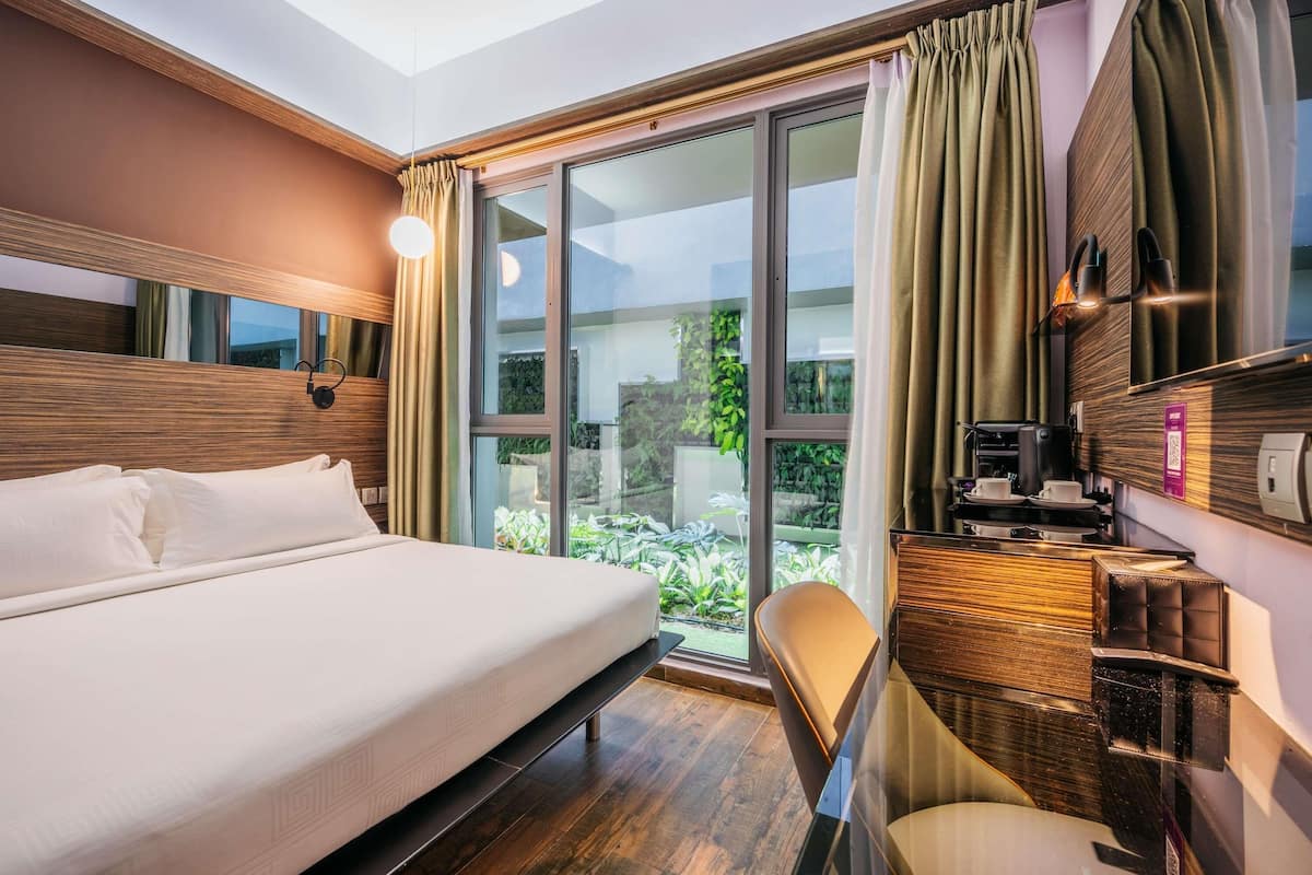 Une chambre d’hôtel avec un grand lit, un bureau équipé d’une machine à café et une vue sur la verdure par les fenêtres.