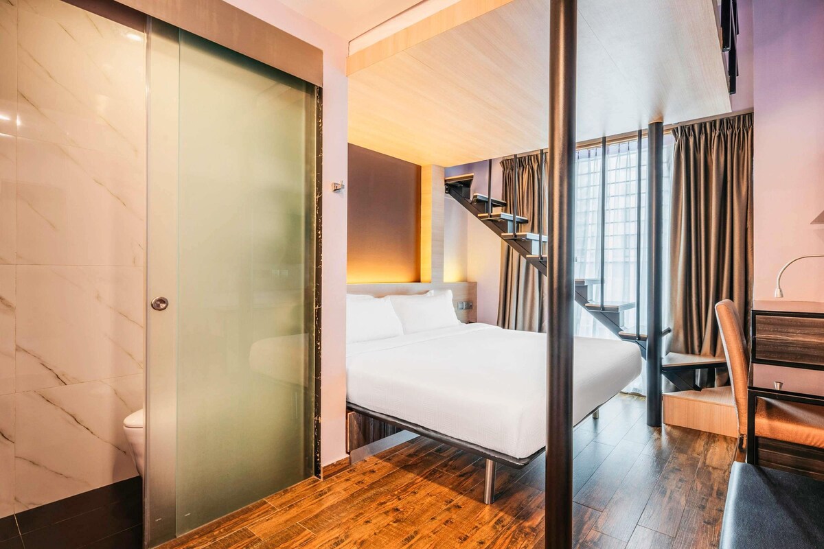 Habitación de hotel con cama, escritorio, silla, escalera y vistas al exterior.